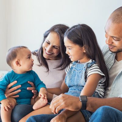 Multiethnic family playing with happy baby son at home. Parent and children relaxing together on the sofa at home in the living room. Little girl sitting on leg of dad looking her new cute brother.